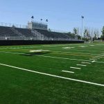 A football field with artificial turf and empty bleachers under a clear blue sky