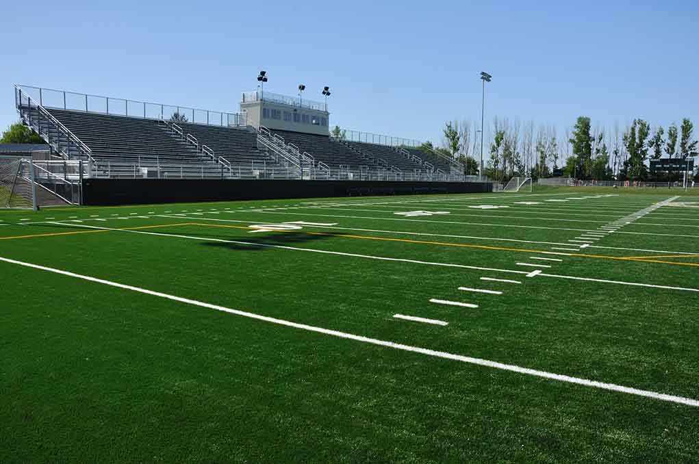 A football field with artificial turf and empty bleachers under a clear blue sky