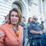 Pelosi Erupts Over Direct Question—Washington Stunned A woman in an orange blazer speaking passionately at a rally with security personnel in the background