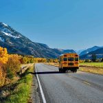 A yellow school bus driving on a rural road surrounded by autumn trees and mountains