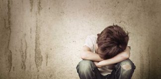 A sad child sitting on the floor with arms wrapped around knees against a concrete wall