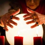 A person with adorned hands hovering over a glowing crystal ball surrounded by candles