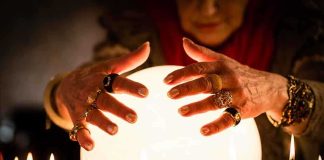 A person with adorned hands hovering over a glowing crystal ball surrounded by candles