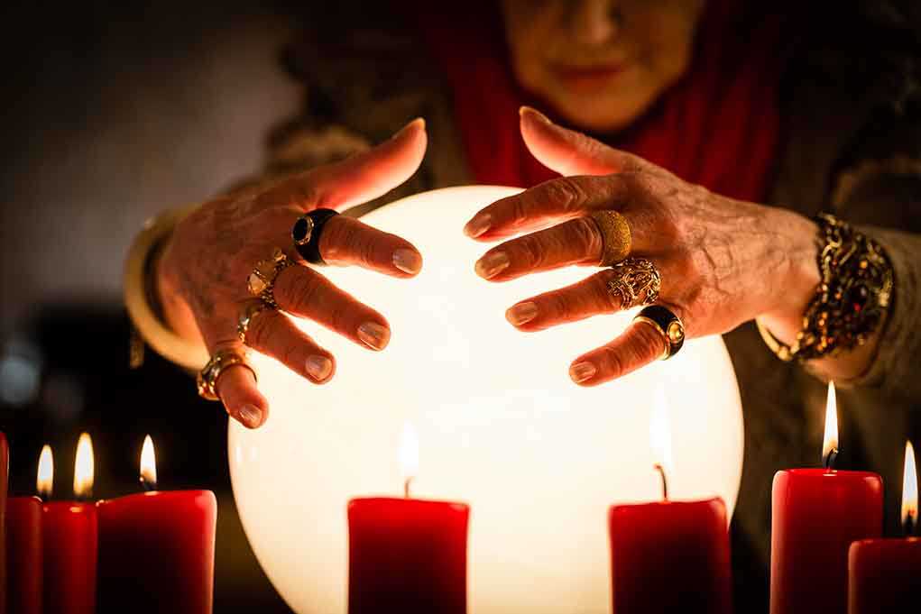 A person with adorned hands hovering over a glowing crystal ball surrounded by candles