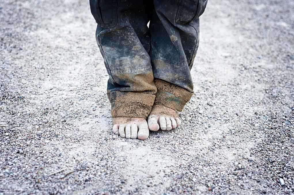 A child standing barefoot on a gravel surface with dirty pants