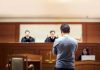 Man stands in courtroom before judges and stenographer.