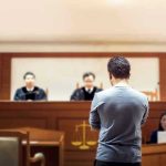Man stands in courtroom before judges and stenographer.