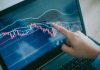 Hand pointing at stock market graph on laptop screen.