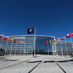 U.S. Considers Expanding Nuclear Reach Flags outside NATO headquarters building under clear blue sky.