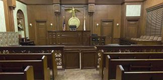 Kill Threat Ignites DOJ Minefield Interior view of an empty courtroom with wooden benches and a judges bench
