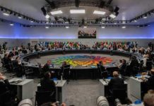 A large round table meeting with global leaders and flags in the background