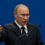 A man in a suit gesturing while speaking at a podium with microphones against a blue background