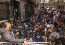 A large group of people sitting on a city street, some wearing masks
