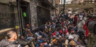 A large group of people sitting on a city street, some wearing masks