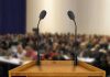 A wooden podium with two microphones in front of a blurred audience