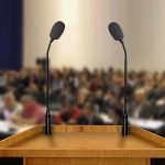 “Suicidal Empathy” Debate Sparks Warning Over Western Decline A wooden podium with two microphones in front of a blurred audience