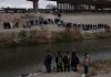 Large group of people lined up on a riverbank below a border fence