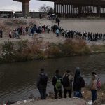 Large group of people lined up on a riverbank below a border fence