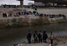 Large group of people lined up on a riverbank below a border fence