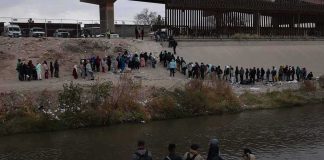 Large group of people lined up on a riverbank below a border fence
