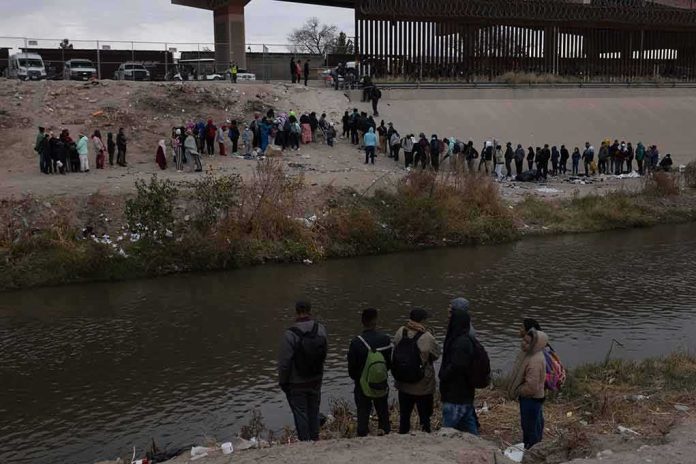Large group of people lined up on a riverbank below a border fence
