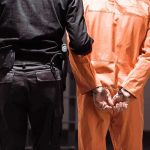 Prisoner in orange jumpsuit handcuffed with guard standing beside
