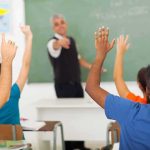 Students raising hands in a classroom while teacher points