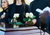 Mourners holding white roses beside a wooden casket