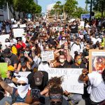 J. C. Tretter, Teddy Williams, and Eugene Sims among protesters kneeling with signs at a large street demonstration