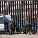 ICE Arrests Surge to New Monthly Record Border agents escort a group near a tall fence and a patrol van