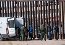 Border agents escort a group near a tall fence and a patrol van