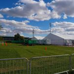 Temporary white tents and equipment on a grassy field with traffic cones and metal barriers