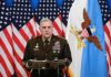 Military officer speaks at podium with U.S. flags behind