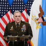 Military officer speaks at podium with U.S. flags behind