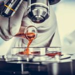 Gloved hand pouring orange liquid into a petri dish under a microscope