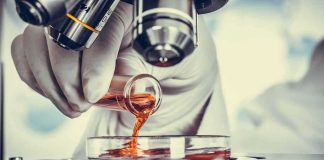 Gloved hand pouring orange liquid into a petri dish under a microscope