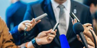 $1.7M Podcast Payment Trail Examined Man in suit surrounded by microphones at press conference