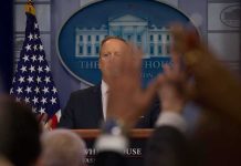 Press secretary at podium with raised hands in briefing room