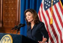 Kathy Hochul speaking at a podium with microphones and a U.S. flag behind her