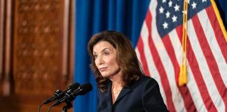 Kathy Hochul speaking at a podium with microphones and a U.S. flag behind her