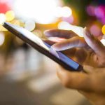 A person's hand using a smartphone in a city at night with blurred lights in the background