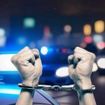 Handcuffed hands raised, police cars with lights on.