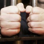 Hands gripping jail cell bars tightly.