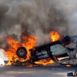 An overturned car engulfed in flames and smoke on a city street