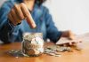 Person placing a coin into a savings jar labeled SAVINGS