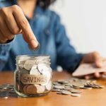 Person placing a coin into a savings jar labeled SAVINGS